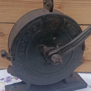 May include: A vintage black cast iron Spong & Co. food mill featuring a circular housing with embossed lettering, a side-mounted hand crank, and a sturdy rectangular base. The metal surface shows signs of age and wear, with a prominent handle.