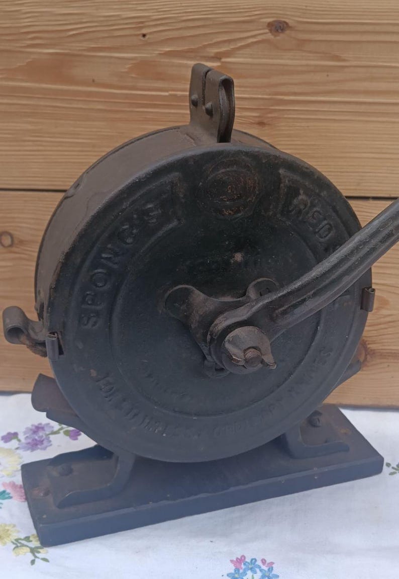 May include: A vintage black cast iron Spong & Co. food mill featuring a circular housing with embossed lettering, a side-mounted hand crank, and a sturdy rectangular base. The metal surface shows signs of age and wear, with a prominent handle.