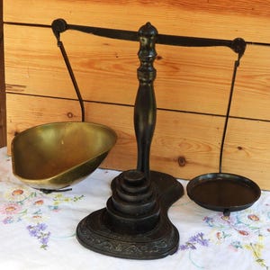 Puede incluir: Una balanza de metal negro con un pilar central sobre una base escalonada, una barra horizontal y dos platos colgantes. Un plato es un cuenco profundo de latón pulido, mientras que el otro es una bandeja circular plana de color negro.