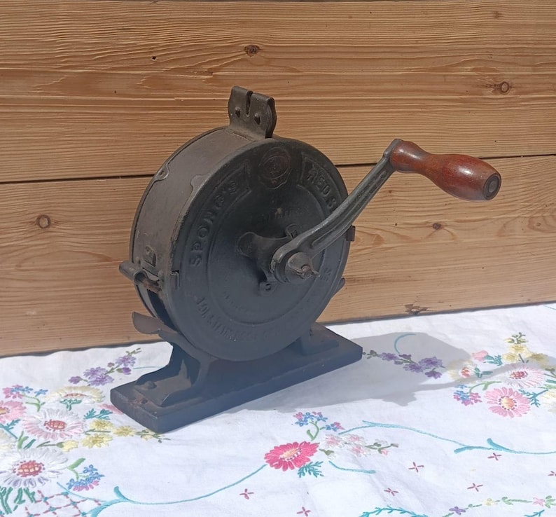 May include: A vintage cast iron kitchen tool with a circular body and a dark grey finish. It features a side-mounted hand crank with a smooth, dark brown wooden handle. The metal casing has embossed lettering and a sturdy base for stability.
