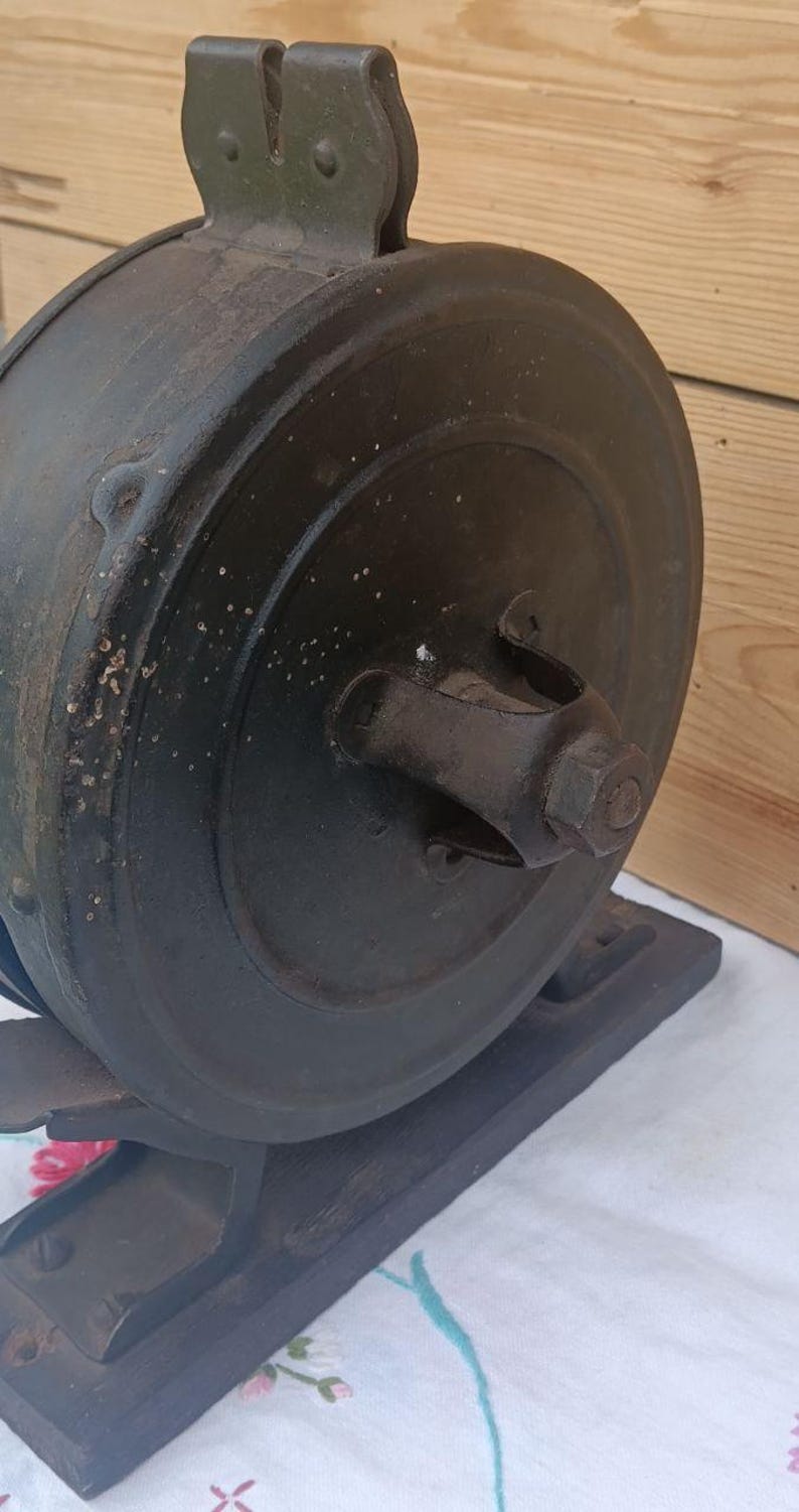 May include: A vintage dark grey metal circular tool with a central hexagonal nut and a top mounting bracket. The object is mounted on a rectangular dark grey metal base, featuring a weathered, textured surface with visible signs of age and wear.