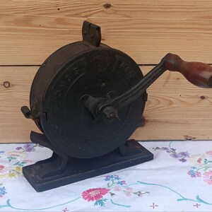 May include: A vintage black cast iron hand-cranked food mill featuring a circular body with embossed lettering and a dark wooden handle. The device is mounted on a rectangular base and includes a metal crank arm for manual operation of the mechanism.