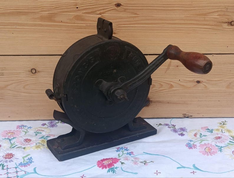 May include: A vintage black cast iron hand-cranked food mill featuring a circular body with embossed lettering and a dark wooden handle. The device is mounted on a rectangular base and includes a metal crank arm for manual operation of the mechanism.