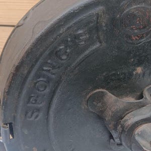 May include: A dark grey cast iron kitchen tool with a circular body featuring the embossed text SPONG'S. The device includes a central rotating mechanism and a top mounting bracket, showing signs of age and wear on the metallic surface.
