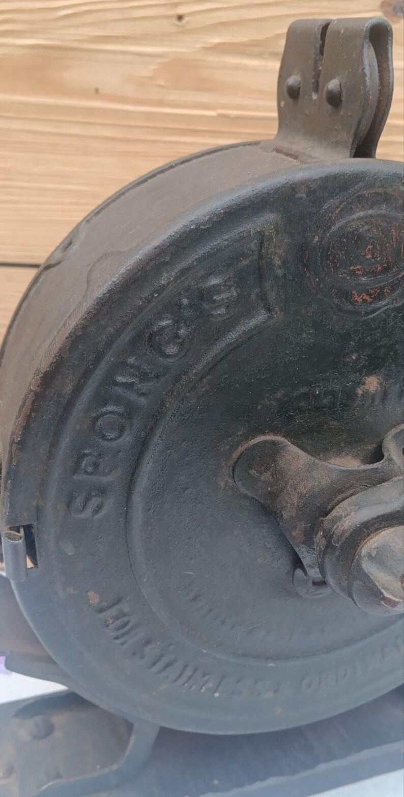 May include: A dark grey cast iron kitchen tool with a circular body featuring the embossed text SPONG'S. The device includes a central rotating mechanism and a top mounting bracket, showing signs of age and wear on the metallic surface.