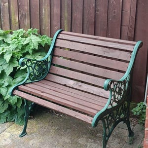 May include: A garden bench featuring horizontal brown wooden slats for the seat and backrest, supported by ornate, scroll-patterned green metal side frames with integrated armrests and legs, providing a sturdy structure for outdoor seating.