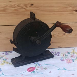 May include: A black cast iron hand-cranked tool with a circular housing and a brown wooden handle. The device features a sturdy rectangular base and a metal crank arm. Embossed lettering is visible on the circular side panel of the mechanism.