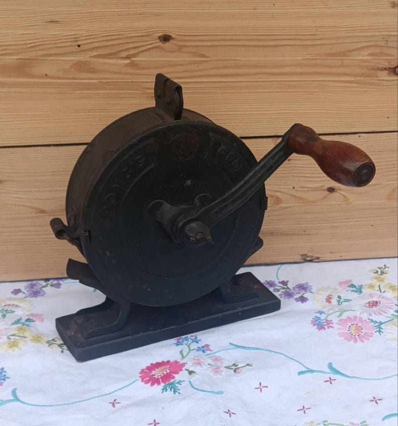 May include: A black cast iron hand-cranked tool with a circular housing and a brown wooden handle. The device features a sturdy rectangular base and a metal crank arm. Embossed lettering is visible on the circular side panel of the mechanism.