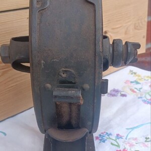 May include: A vintage cast iron hand-cranked tool with a dark, weathered patina. The device features a central circular housing, a protruding horizontal axle with a nut, and a vertical base. A small metal chute extends from the front of the unit.