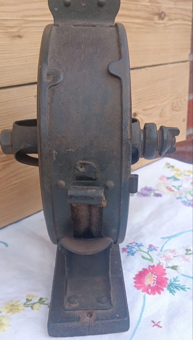 May include: A vintage cast iron hand-cranked tool with a dark, weathered patina. The device features a central circular housing, a protruding horizontal axle with a nut, and a vertical base. A small metal chute extends from the front of the unit.