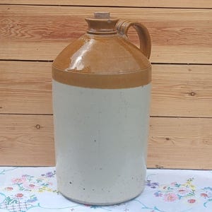 May include: A tall, cylindrical stoneware jug featuring a two-tone glaze. The upper portion and handle are finished in a glossy, warm brown, while the lower body is a matte, off-white cream color. A small, light-colored cork stopper sits in the top opening.
