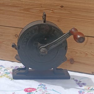 May include: A vintage cast iron Spong's kitchen mill featuring a circular body with embossed lettering, a central hand crank, and a smooth brown wooden handle. The device is mounted on a rectangular base and includes a small metal lever on the side.
