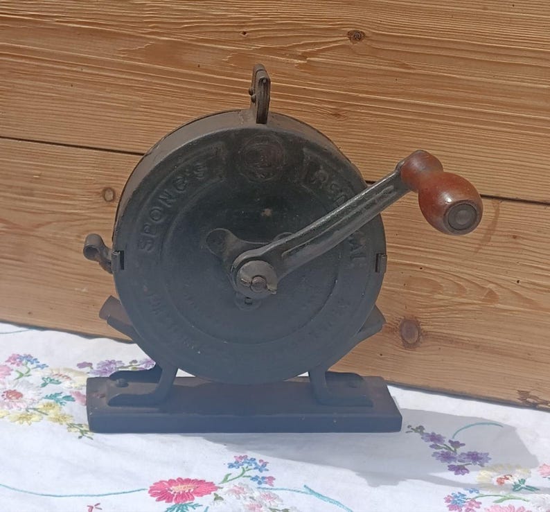 May include: A vintage cast iron Spong's kitchen mill featuring a circular body with embossed lettering, a central hand crank, and a smooth brown wooden handle. The device is mounted on a rectangular base and includes a small metal lever on the side.