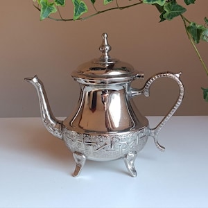 Może przedstawiać: Srebrny czajnik z ozdobnym wzorem i trzema nóżkami. Czajnik ma pokrywkę z gałką na górze.