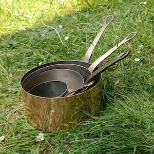 May include: A set of four copper-colored saucepans with long, riveted handles. The pans are nested inside each other, with the largest pan at the bottom. The pans are sitting in green grass.