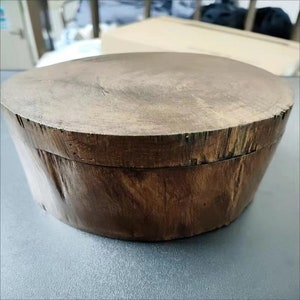 May include: A round wooden box with a lid. The box is made of dark brown wood with a natural wood grain. The lid is slightly raised and has a smooth surface.