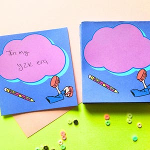 Puede incluir: Dos blocs de notas cuadrados azules con un diseño de nube rosa y el texto "In my y2k era". Cada bloc de notas presenta un lápiz y las piernas y los pies de una persona. Hay cuentas de colores esparcidas.