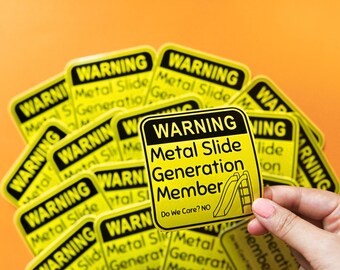 Puede incluir: Pegatinas amarillas con texto negro que dice "WARNING Metal Slide Generation Member" y un gráfico de tobogán. Las pegatinas están sobre un fondo naranja. Una pegatina está siendo sostenida.