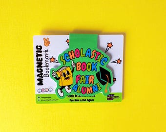 Puede incluir: Un marcapáginas magnético con un diseño verde y blanco. El marcapáginas presenta el texto "SCHOLASTIC BOOK FAIR ALUMNI" y un personaje de libro de dibujos animados. El fondo es amarillo.