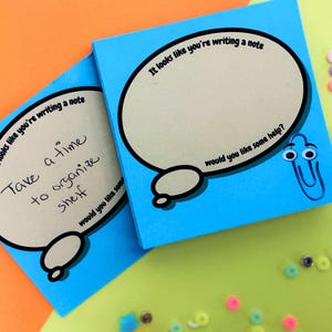 May include: Two blue sticky note pads with speech bubble designs. One pad has handwritten text: "Take a time to organize shelf." The other pad has the text: "It looks like you're writing a note. Would you like some help?" A paperclip with googly eyes is on one pad.