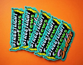 Puede incluir: Un montón de pegatinas con un borde turquesa y las palabras "Streetlight Curfews" en blanco, verde y negro. Un rayo amarillo en la parte superior. La palabra "Baddies" está en naranja y rosa.