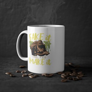 Pode incluir: Caneca de café de cerâmica branca com um gráfico amarelo que diz "Fake it till you make it". O gráfico também inclui uma imagem de desenho animado de um gorila sentado em frente a palmeiras.
