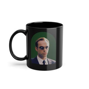 Puede incluir: Taza de café negra con borde y asa blancos. La taza presenta un gráfico de un hombre con gafas de sol sobre un fondo verde.