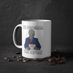 Puede incluir: Taza de cerámica blanca con una ilustración en negro y azul de una persona sosteniendo un cartel que dice "NPC". El texto "WE PROGRAM YOU REPEAT" está impreso en la taza.