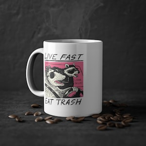 Op de afbeelding: Een witte keramische mok met een zwart-witte illustratie van twee wasberen die een auto besturen. De tekst "LIVE FAST EAT TRASH" staat op de mok gedrukt. De wasberen dragen een zonnebril en degene die achter het stuur zit heeft de tekst "LIVING THE LIFE!" erboven gedrukt.