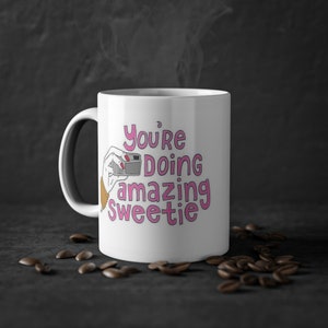 Könnte beinhalten: Weiße Keramiktasse mit einem pinkfarbenen und schwarzen Design. Die Tasse zeigt eine Hand, die eine Kamera hält, und den Text "You're doing amazing sweetie".