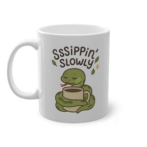 Könnte beinhalten: Weiße Keramik-Tasse mit einem Cartoon-Schlangen-Design. Die Tasse zeigt den Text "SssIPPIN' SLOWLY" mit grünen Blättern und einem goldenen Stern. Die Schlange ist um eine Kaffeetasse gewickelt.