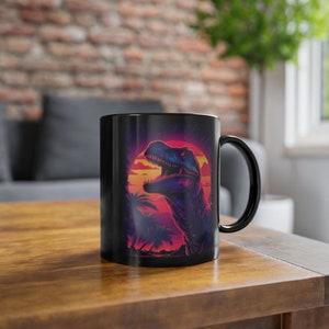 Op de afbeelding: Zwarte keramische mok met een afbeelding van een dinosaurus in een retro zonsondergangsscene. De dinosaurus kijkt naar rechts en heeft een fel roze en oranje zonsondergang achter zich.