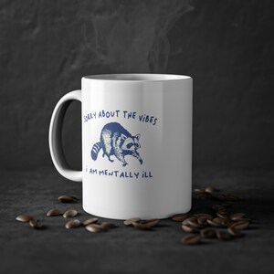 White ceramic mug with funny raccoon meme and caption Sorry About the Vibes, I Am Mentally Ill