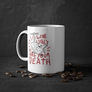 Peut inclure: Une tasse à café en céramique blanche avec une illustration en noir et blanc d'un loup avec le texte "Live Ugly Take Your Death" en rouge.