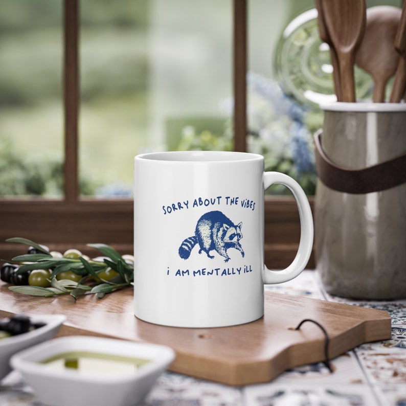 White ceramic mug with funny raccoon meme and caption Sorry About the Vibes, I Am Mentally Ill