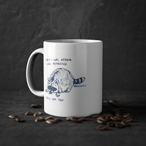 Può includere: Una tazza in ceramica bianca con un'illustrazione in bianco e nero di un procione. Il testo sulla tazza recita "First I was afraid. I was petrified. Still am tbh. Raccdaily."