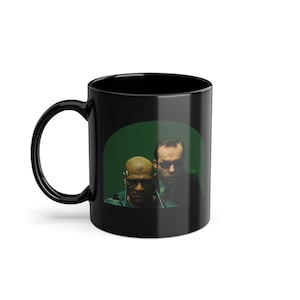 Puede incluir: Taza de café de cerámica negra con un fondo verde que presenta dos caras de hombres. Los hombres están mirando a la cámara. La taza es un gran regalo para los fanáticos de Matrix.