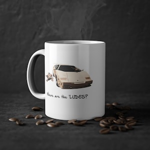 May include: White ceramic mug with a beige sports car graphic and the text "Where are the 'LUDES'?" The mug has a black handle and is surrounded by coffee beans. A fun gift for car enthusiasts.
