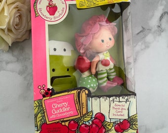 Może przedstawiać: Lalka Cherry Cuddler Strawberry Shortcake z zwierzątkiem Gooseberry. Lalka ma na sobie różową sukienkę w zielone kropki i zielono-białą w paski koszulę. Lalka trzyma agrest. Lalka i zwierzak pachną wiśnią. W pudełku znajduje się kartka z podziękowaniem.