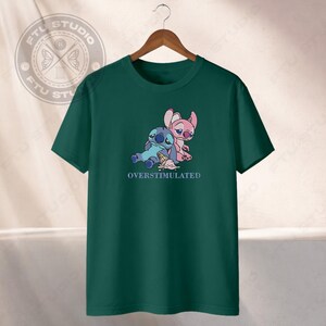 May include: A dark green t-shirt featuring a cartoon illustration of two characters, one blue and one pink, with an ice cream cone. The text "OVERSTIMULATED" is below the characters.