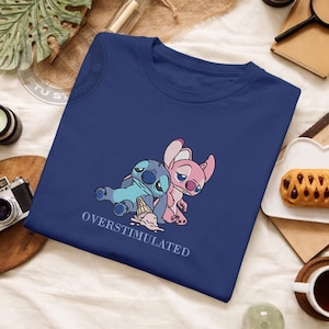 May include: A navy blue t-shirt featuring an embroidered design of two cartoon characters, one blue and one pink, with the text "OVERSTIMULATED" below. The blue character is lying down with a spilled ice cream cone.