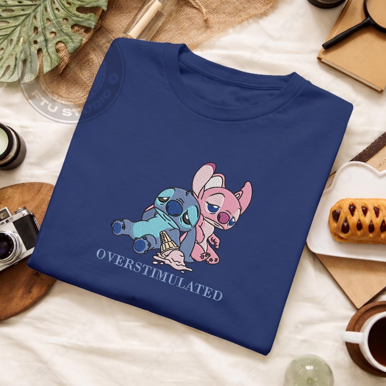 May include: A navy blue t-shirt featuring an embroidered design of two cartoon characters, one blue and one pink, with the text "OVERSTIMULATED" below. The blue character is lying down with a spilled ice cream cone.