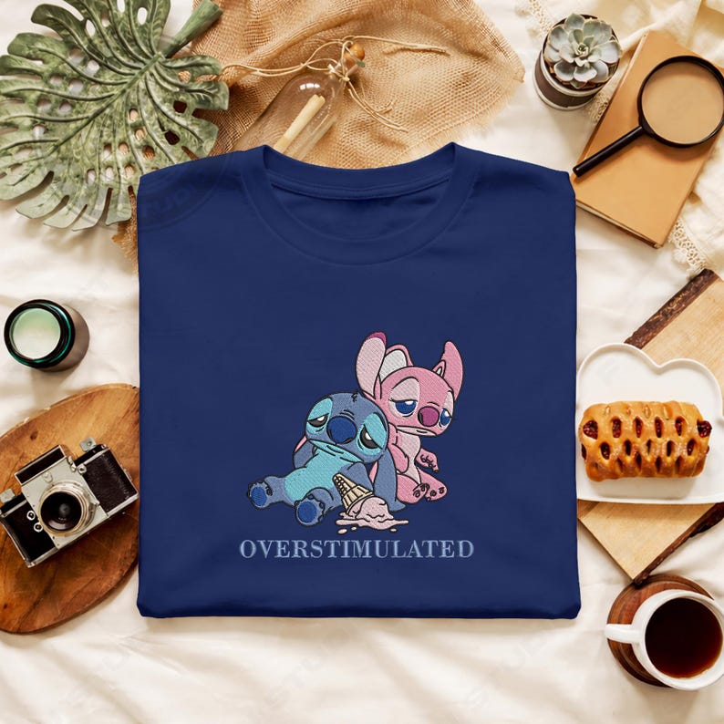 May include: Navy blue t-shirt featuring embroidered cartoon characters Stitch and Angel, with a melting ice cream cone. The text "OVERSTIMULATED" is below the characters. The shirt is folded on a white surface.