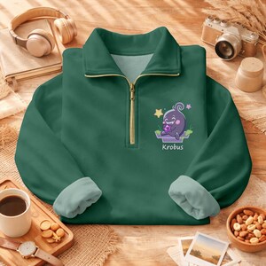May include: A dark green pullover sweatshirt with a quarter-zip collar and light green cuffs. The sweatshirt features a cartoon graphic of a purple creature with the name "Krobus" below it. The sweatshirt is on a wooden surface.