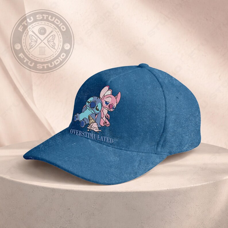 May include: A blue baseball cap featuring a cartoon image of two characters, one blue and one pink, with the text "OVERSTIMULATED". The cap has a curved brim and a structured crown. The design is on the front of the cap.