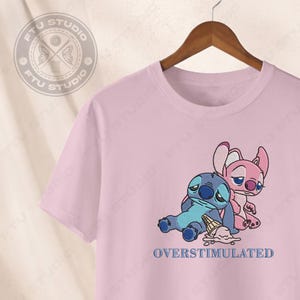 May include: A light pink t-shirt featuring an embroidered design of two cartoon characters, one blue and one pink, with a spilled ice cream cone. The text "OVERSTIMULATED" is below the characters.