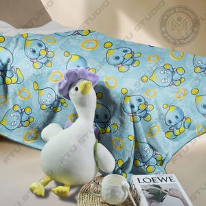 May include: A blue and yellow fleece blanket with a repeating pattern of a blue character with yellow rings. A white plush duck with a purple flower on its head is sitting on the blanket. A white mug and a book with the word "LOEWE" on the cover are in the foreground.