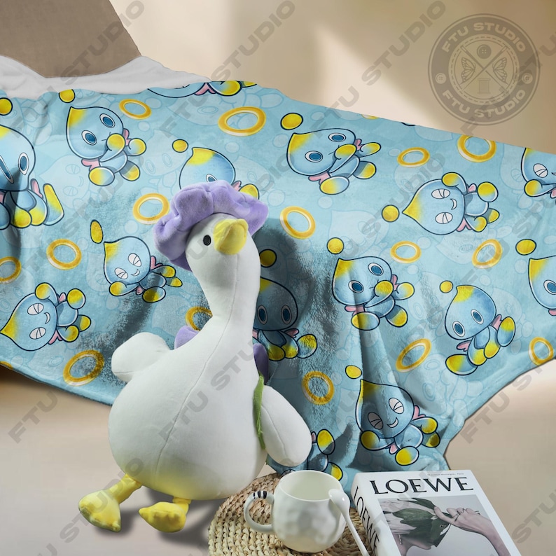 May include: A blue and yellow fleece blanket with a repeating pattern of a blue character with yellow rings. A white plush duck with a purple flower on its head is sitting on the blanket. A white mug and a book with the word "LOEWE" on the cover are in the foreground.