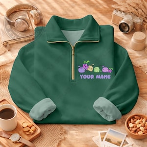 Könnte beinhalten: Ein dunkelgrünes Pullover-Sweatshirt mit einem Viertel-Reißverschlusskragen und einem hellgrünen Innenfutter. Das Sweatshirt zeigt ein Cartoon-Design von mehreren kleinen, bunten Figuren und den Text "YOUR NAME" in Weiß.