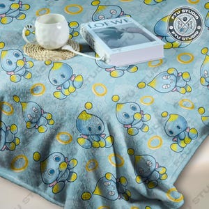 May include: A blue and yellow fleece blanket featuring a repeating pattern of a blue character with yellow accents and gold rings. The blanket is soft and cosy and ideal for snuggling up on the sofa.